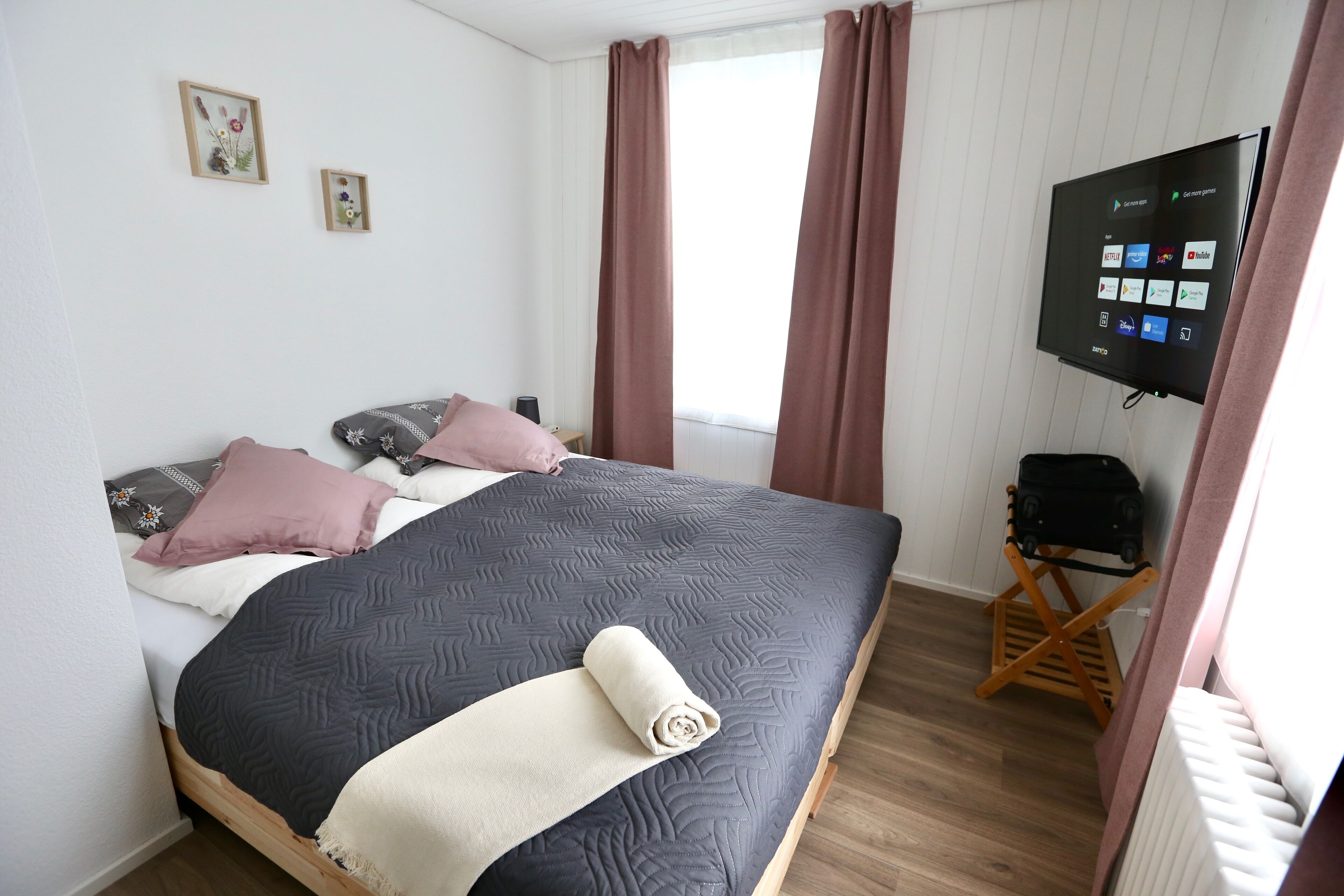 En pænt redt seng med et mørkt dynebetræk, et sammenrullet hvidt tæppe og flere puder. Et fladskærms-tv er monteret på væggen og viser en skærm med forskellige ikoner.