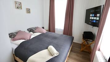 En pænt redt seng med et mørkt dynebetræk, et sammenrullet hvidt tæppe og flere puder. Et fladskærms-tv er monteret på væggen og viser en skærm med forskellige ikoner.