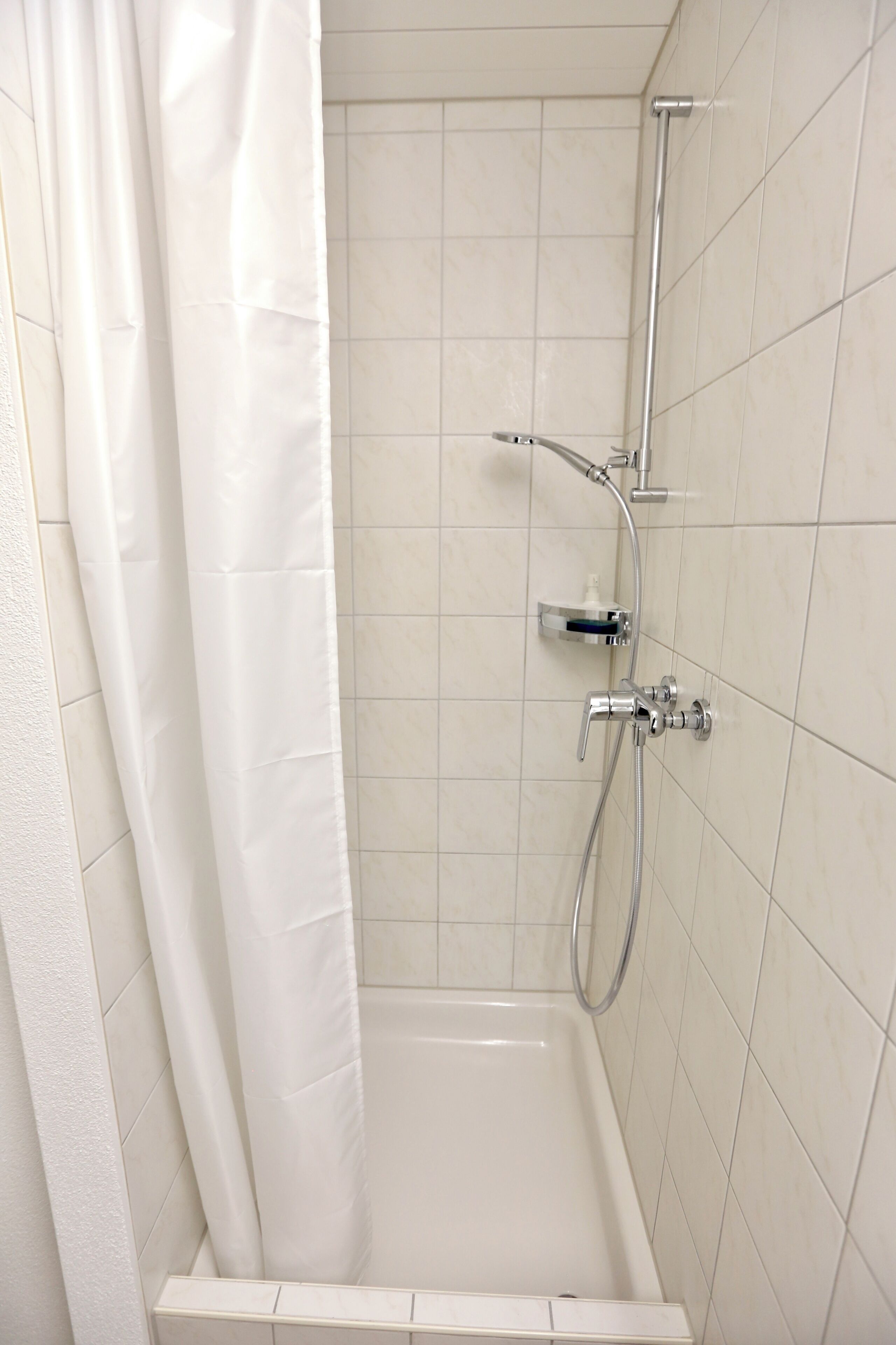Une douche avec un rideau blanc, une douchette à main et un robinet fixé au mur.