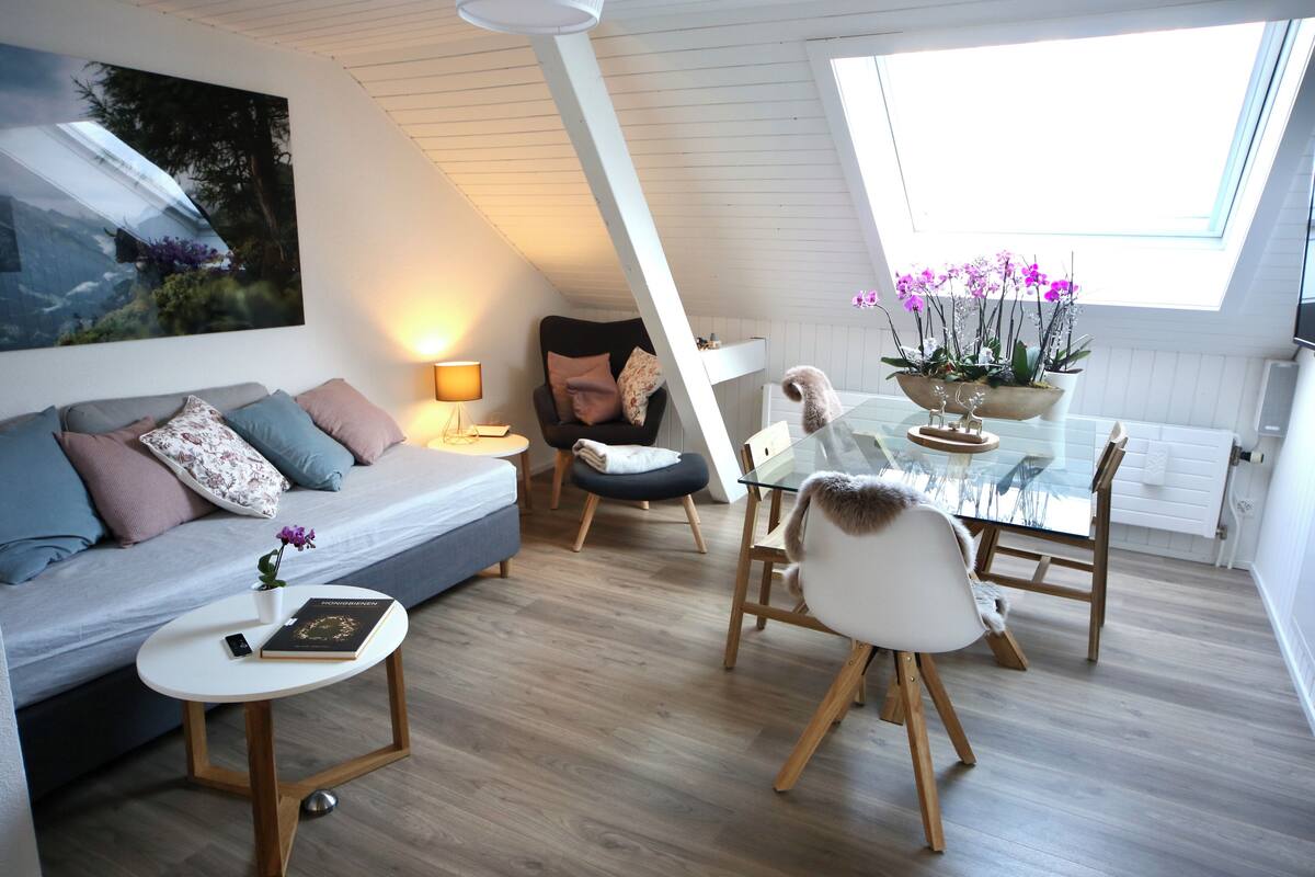 A modern living room with a sofa, a small table, a glass dining table, and a chair. There is a painting on the wall and a window with plants.
