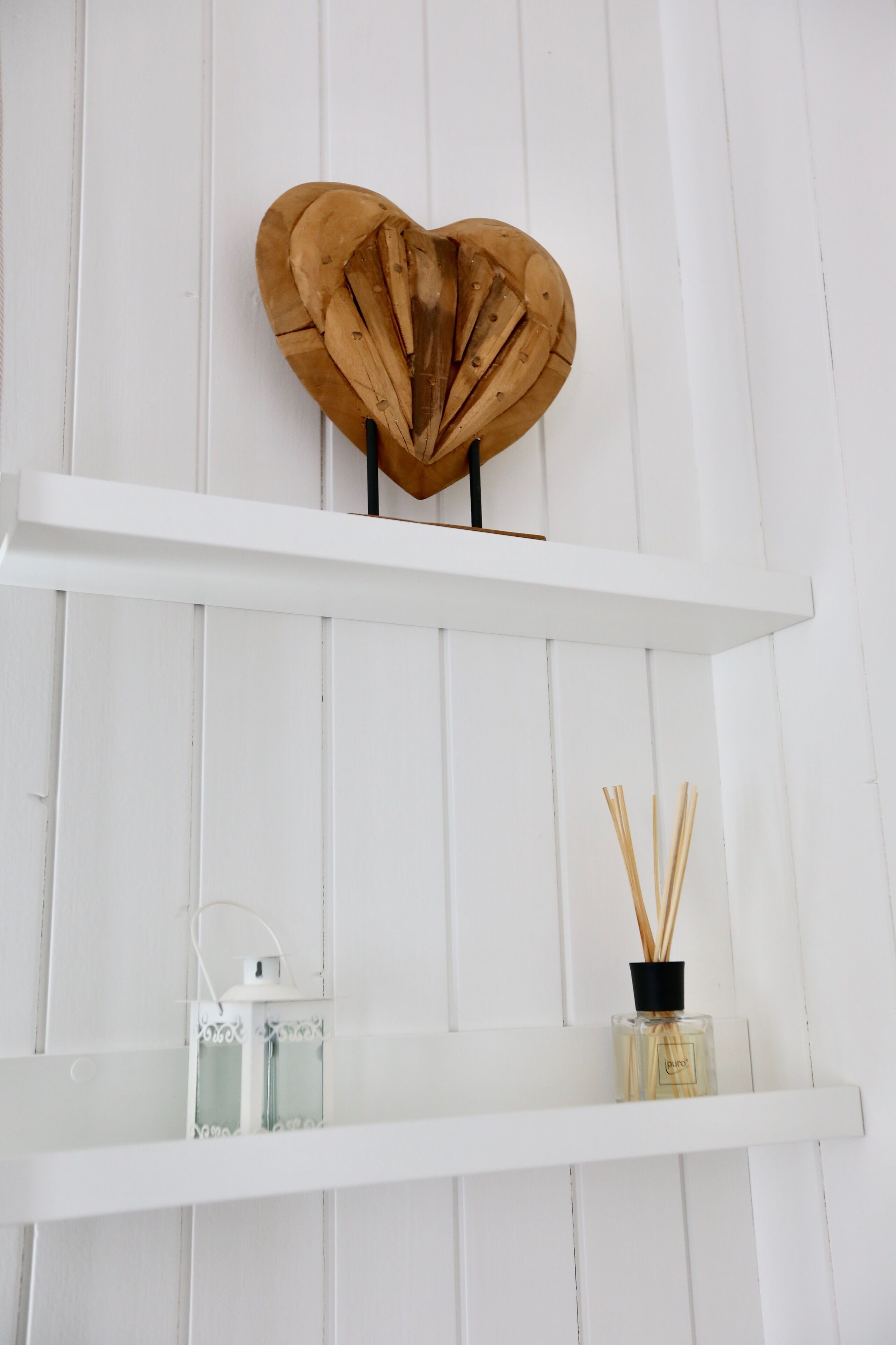 Eine weiße Holzwand mit zwei weißen Regalen. Das obere Regal zeigt ein herzesförmiges Holzobjekt, und das untere Regal beherbergt eine Glasflasche mit schwarzem Verschluss und Rattan-Diffusorstäbchen.