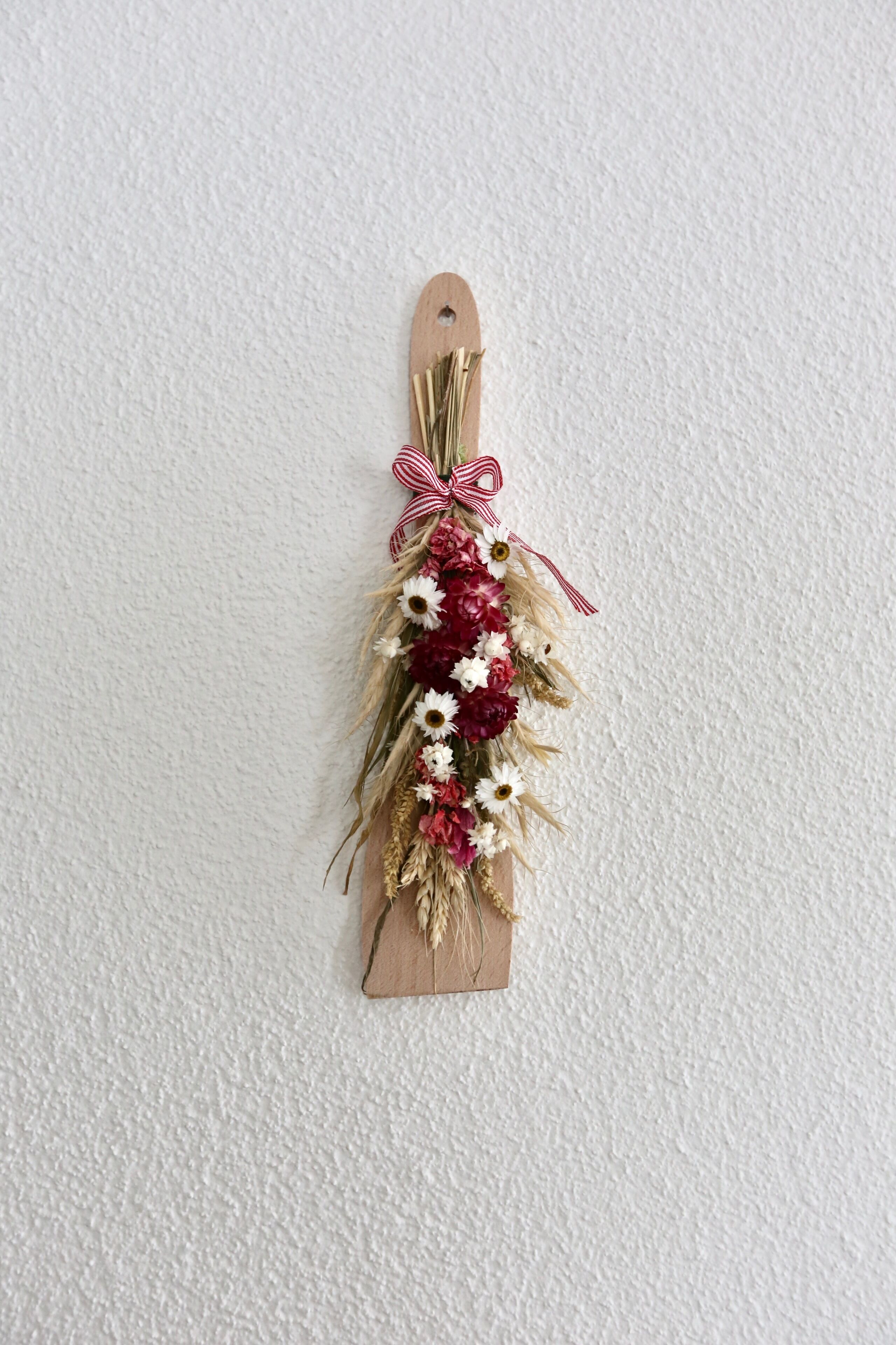 Une plaque en bois ornée d’un ruban rouge et blanc, décorée de fleurs séchées et de verdure.