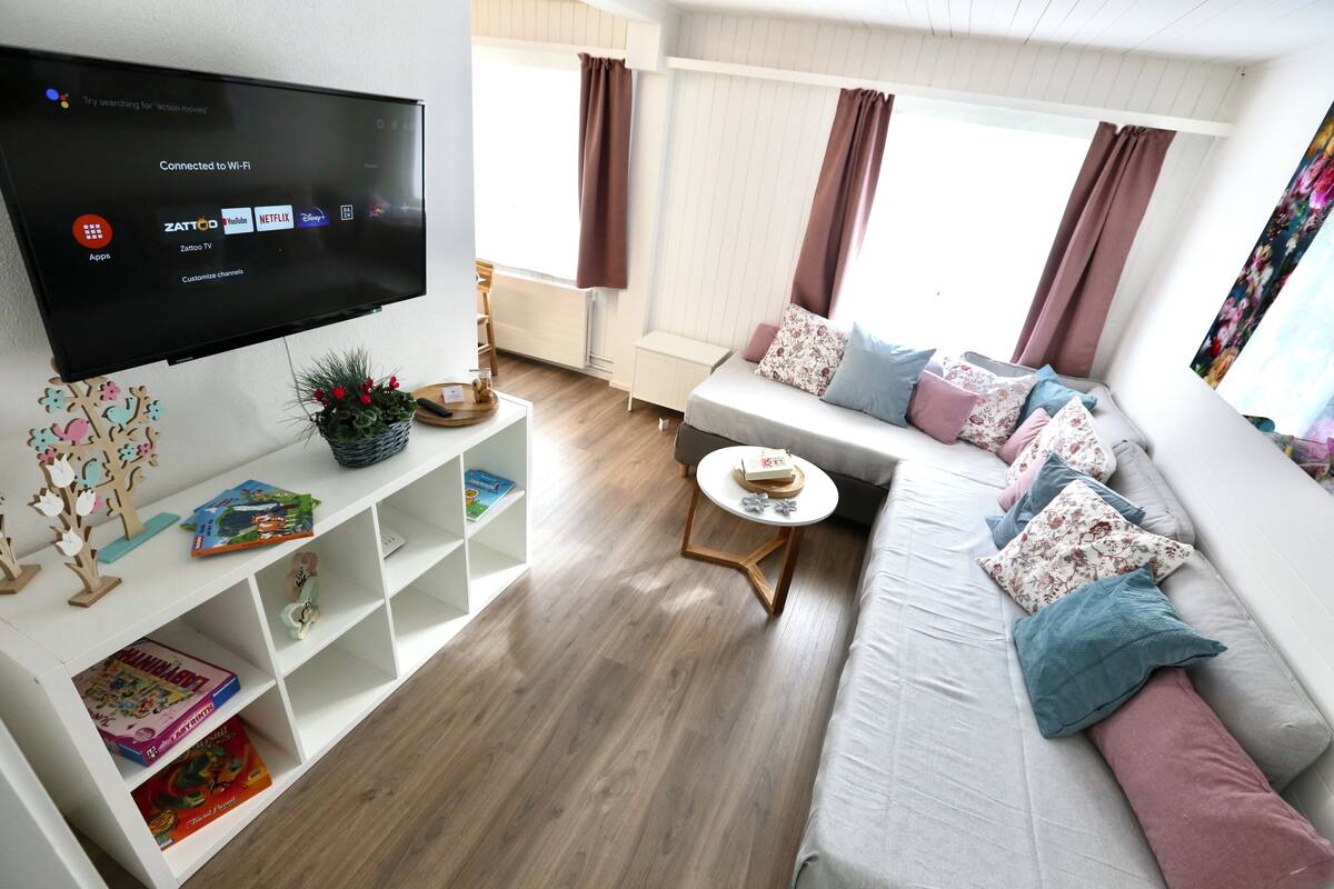 A modern living room with a flat-screen TV, a sofa with cushions, a small round table, and a bookshelf.