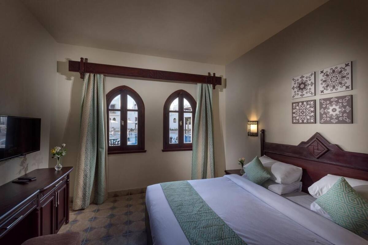 A hotel room with a bed, a TV, a desk, and two windows with curtains.