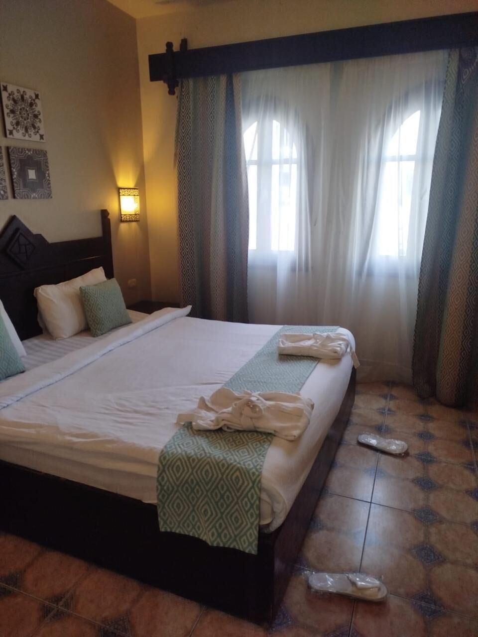 Quarto de hotel com uma cama grande, uma mesa de cabeceira, uma luminária fixada na parede e uma janela com cortinas.
