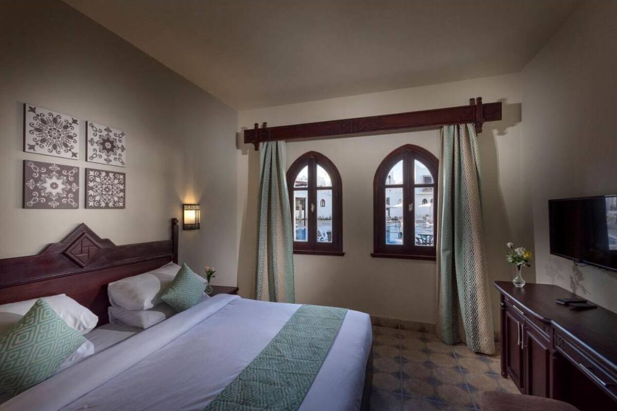 A hotel room with a bed, a TV, a desk, and two windows with curtains.