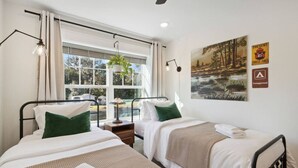 Ein Hotelzimmer mit zwei Betten, einem Fenster mit Vorhängen, einer Lampe, einer Pflanze und einem Poster an der Wand.
