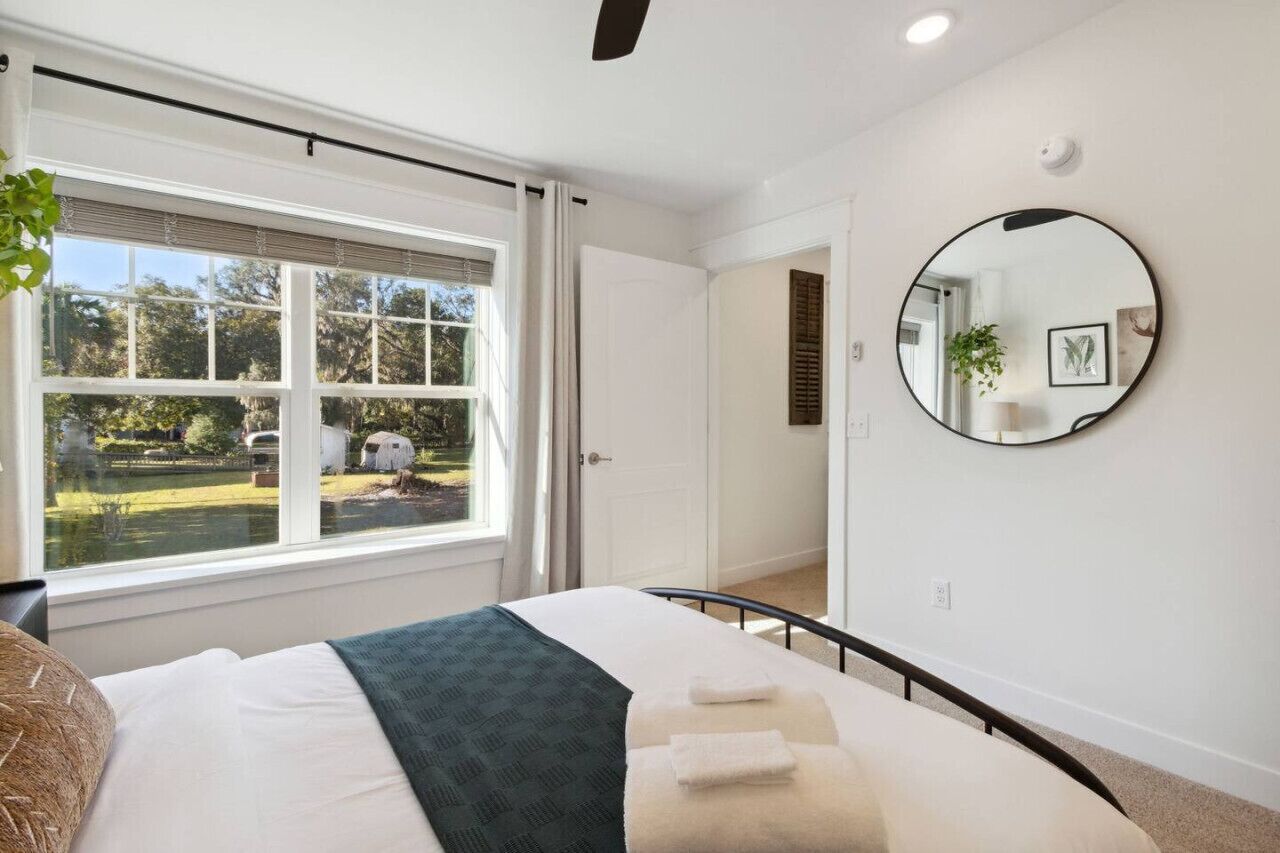 Une chambre avec une grande fenêtre, un lit recouvert d’une couvre-lit verte et blanche, un miroir rond accroché au mur, et une vue sur une cour avec une maison et des arbres.
