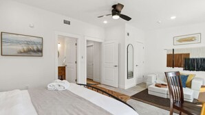 Habitación de hotel con cama, ventilador de techo, baño con ducha, zona de estar con sofá y sillón, y un cuadro en la pared.