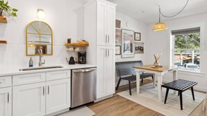 Una cocina moderna con muebles blancos, un lavavajillas de acero inoxidable, una mesa de comedor de madera con un banco y una lámpara colgante.