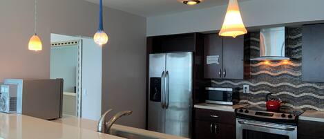 A modern kitchen with a stainless steel refrigerator, microwave, and stove. There are pendant lights above the counter and a backsplash with a mosaic pattern.