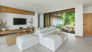 Un salon moderne avec un canapé d’angle, un téléviseur à écran plat, une console en bois et une grande fenêtre donnant sur un jardin.