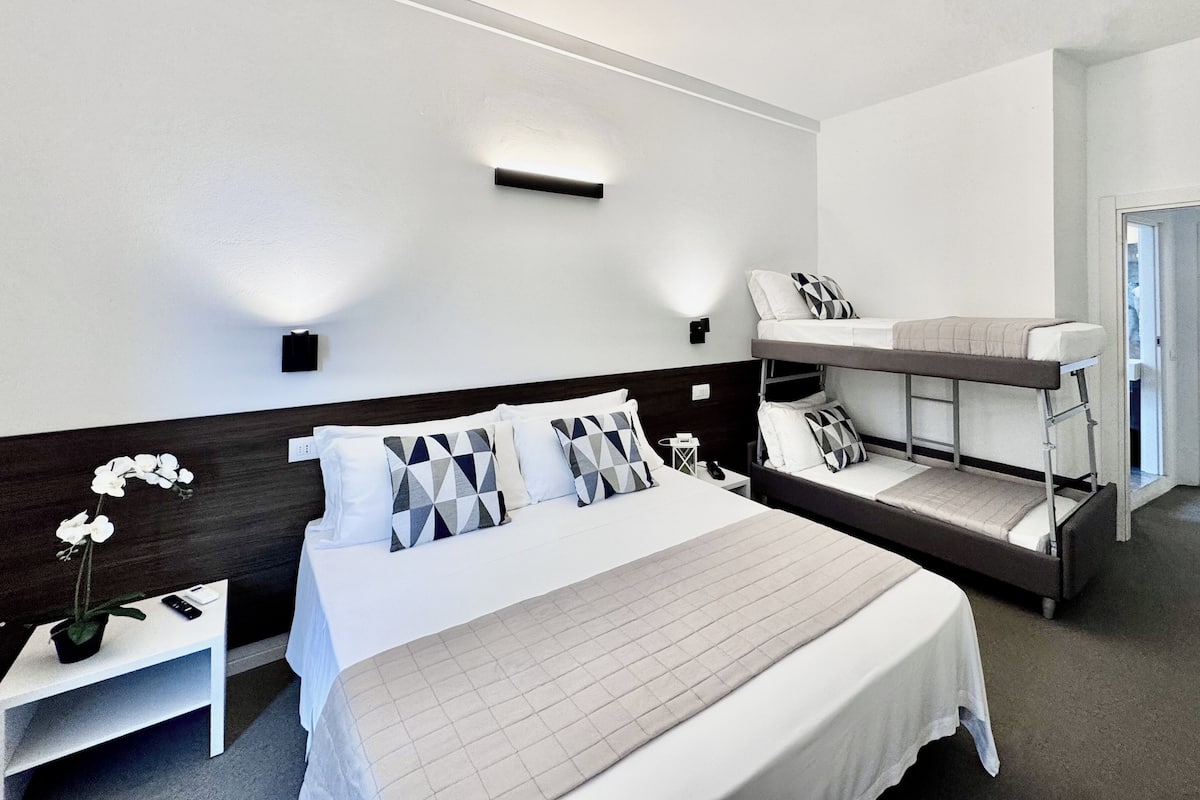 A hotel room with a large bed, a smaller bed on a platform, a bedside table with a phone and a plant, and a wall-mounted light fixture.