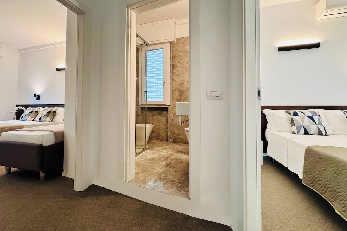 A hotel room with a bed, a seating area, a bathroom with a shower, and a window with blinds.