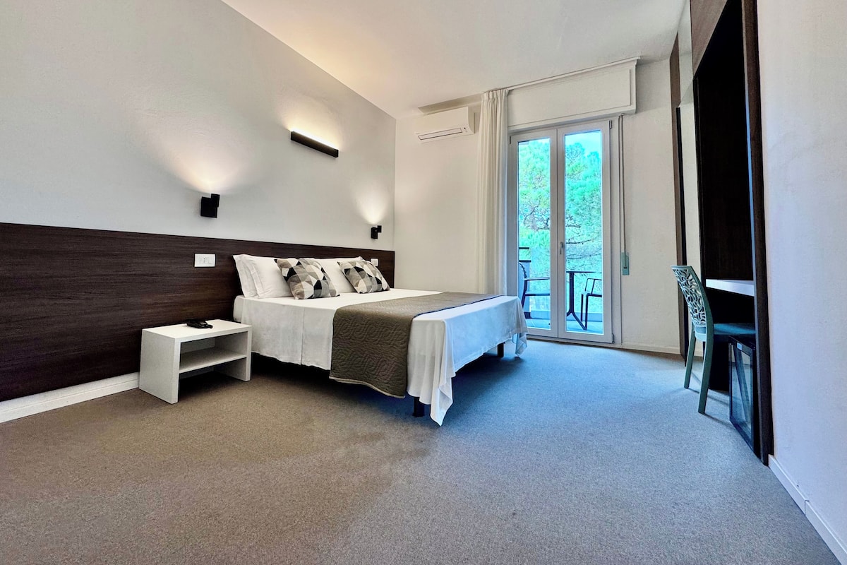 A modern hotel room with a large bed, a bedside table, a small desk, and a chair. There is a sliding glass door leading to an outdoor area.