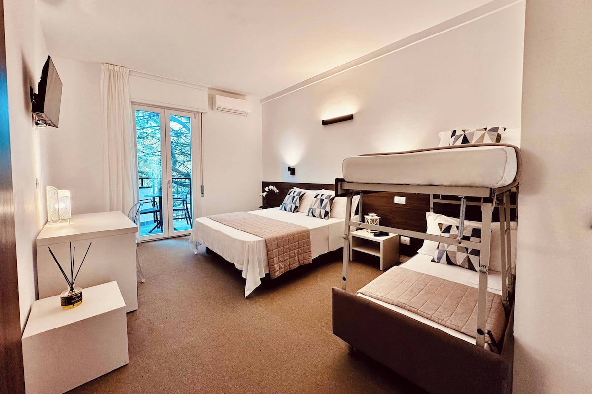 A hotel room with two beds, a small table, a chair, a TV, and a balcony with a view of trees.