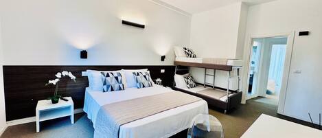 Habitación de hotel moderna con una cama grande, una cama nido, una mesita de noche con lámpara y una silla transparente.