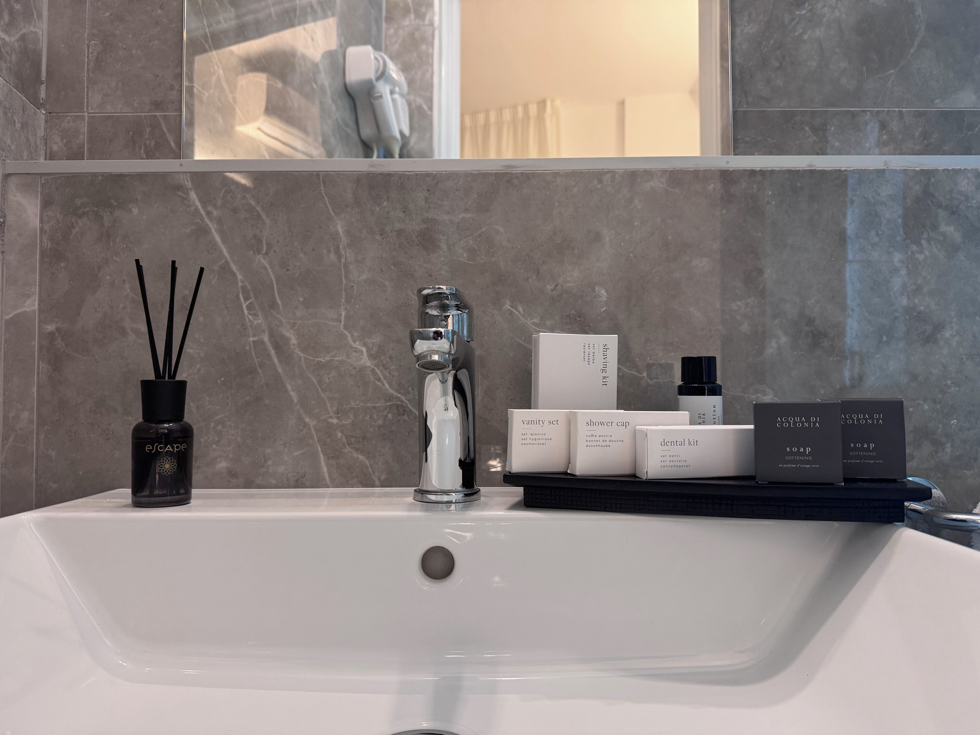Une salle de bain avec un lavabo blanc, un miroir et une étagère contenant divers articles de toilette, notamment un kit de rasage, une trousse de toilette et un flacon de savon.