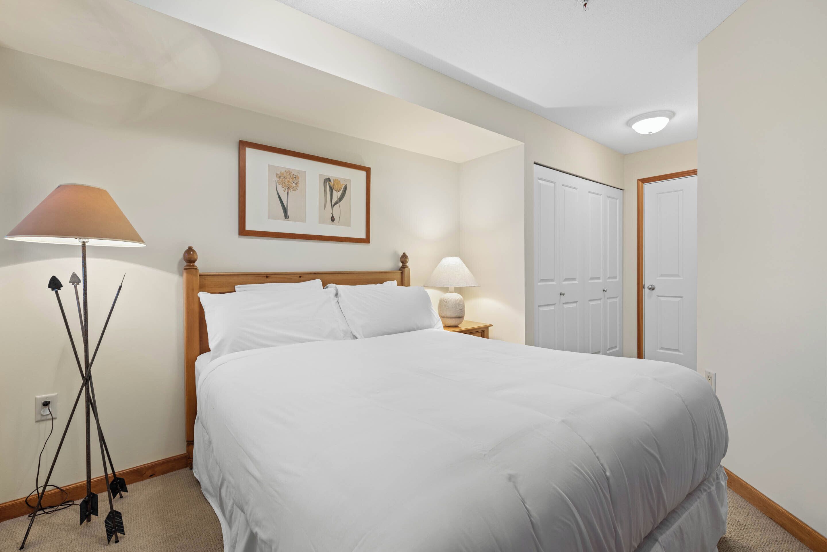A neatly made bed in a hotel room with a headboard, two bedside lamps, a framed floral artwork on the wall, and a door to the right.