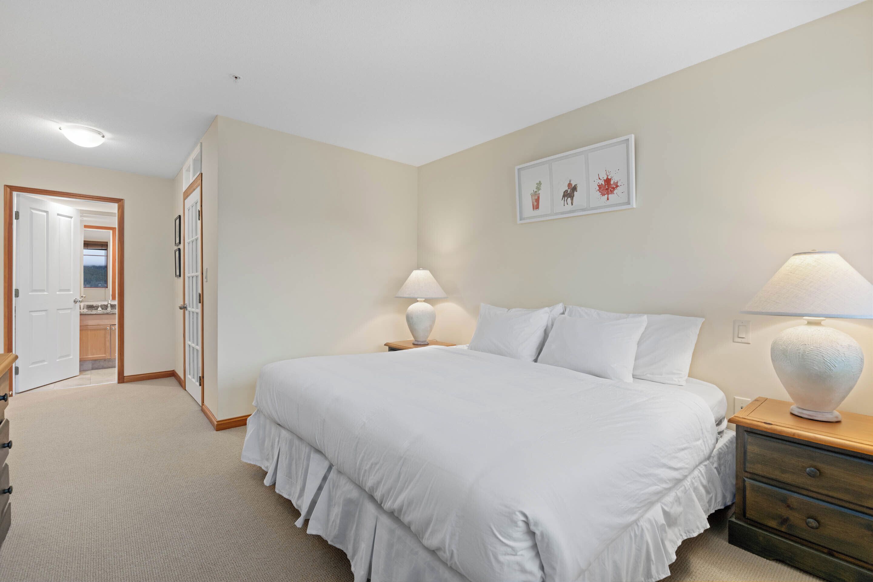 A bedroom with a large bed, two bedside tables with lamps, a door leading to a bathroom, and a framed picture on the wall.