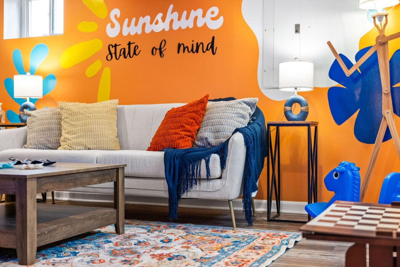 A room with a white sofa, colorful pillows, a patterned rug, a wooden coffee table, a lamp, and a wall with a sunflower mural.