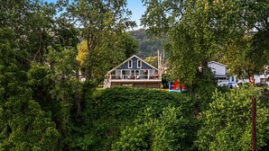 Et hus med stor terrasse, omgivet af frodig vegetation og et vandareal.