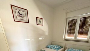 A room with two single beds, each with a floral-patterned bedspread and a pillow. There are two framed pictures on the wall, a window with blinds, and a door to the left. - Seafront Paradise at Rab Island - Emily 3 (Kampor)