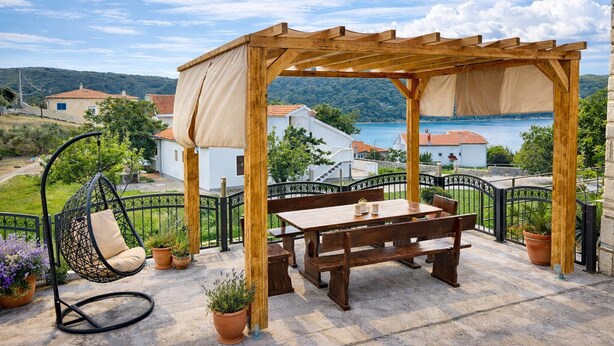 A wooden pergola with a table and chairs, a hanging chair, and potted plants on a terrace with a view of a lake and houses. - Seafront Paradise at Rab Island - Emily 3 (Kampor)
