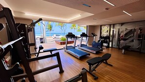A modern gym with treadmills, weight machines, and a bench. It has large windows with a view of the beach and palm trees. - Unity Cabo Branco (João Pessoa)