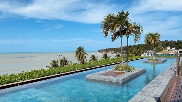A swimming pool with clear blue water, surrounded by palm trees and a view of the beach. - Unity Cabo Branco (João Pessoa)