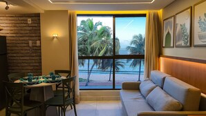 A room with a dining area, a sofa, and a large window with a view of palm trees and the sea. - Unity Cabo Branco (Jo ão Pessoa)