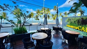 An outdoor seating area with tables and chairs, overlooking a street with cars and palm trees. - Unity Cabo Branco (João Pessoa)