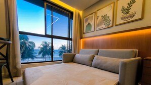 A hotel room with a large bed, a view of palm trees and the ocean through a window, and artwork on the wall. - Unity Cabo Branco (João Pessoa)