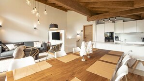 A modern living room with a wooden ceiling, a dining area with a wooden table, and a kitchen in the background. - Superior Ferienhaus # 3B mit Sauna (Uttendorf)