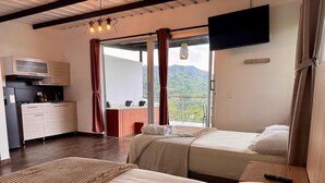 A hotel room with two beds, a kitchenette, a TV, and a balcony with a view of the hills.