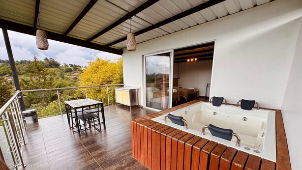 A balcony with a wooden table and chairs, a hot tub, and a glass door leading to a bedroom.