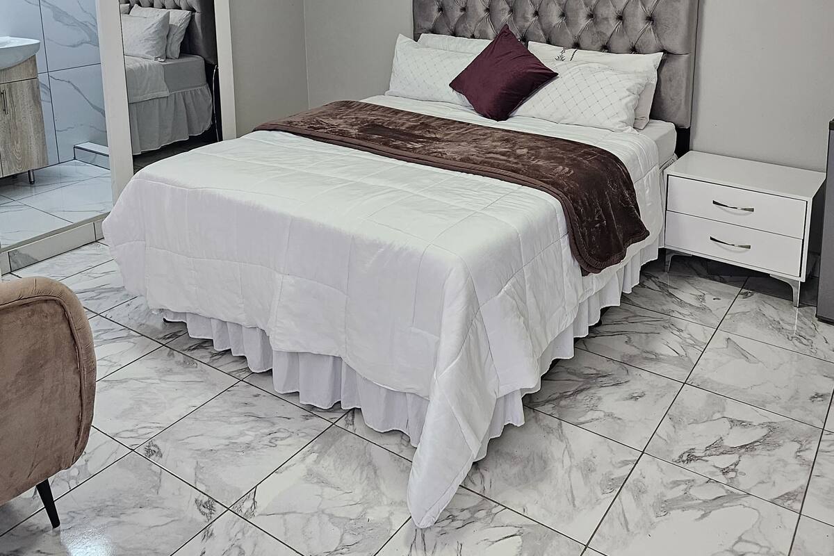 Quarto com uma cama grande, uma mesa de cabeceira, uma cadeira e um banheiro visível por uma porta aberta.