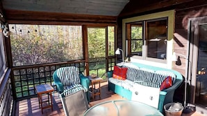 Una cabaña de madera con un porche, un sofá, dos sillones, una mesa y un ventanal con vistas a árboles.