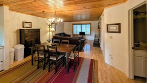 Un área de comedor con techo de madera, chimenea, una mesa de comedor con sillas y una cocina al fondo.