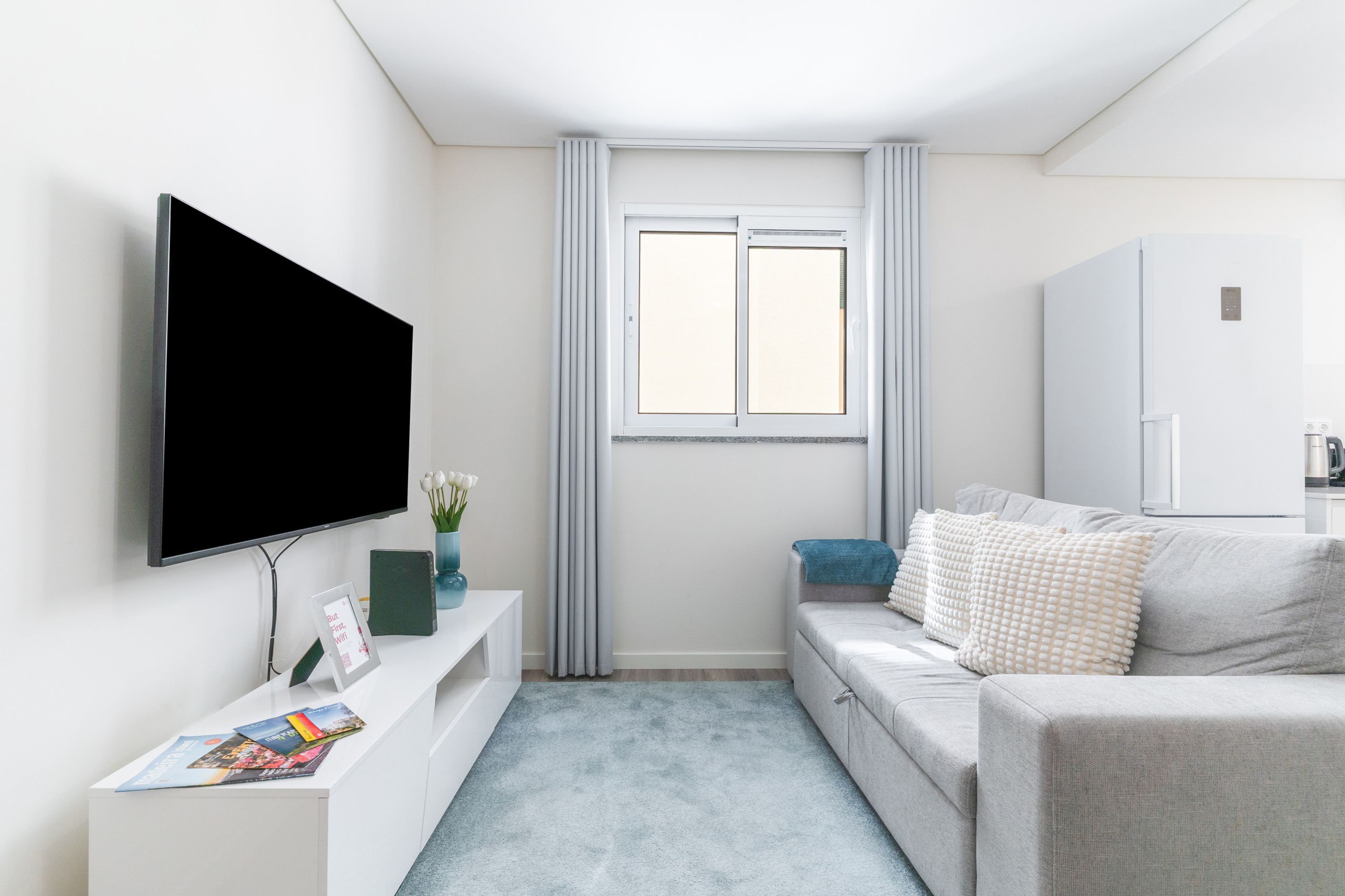 A modern living room with a flat-screen TV mounted on the wall, a sofa with decorative pillows, a small side table with books and a vase, and a window with curtains.