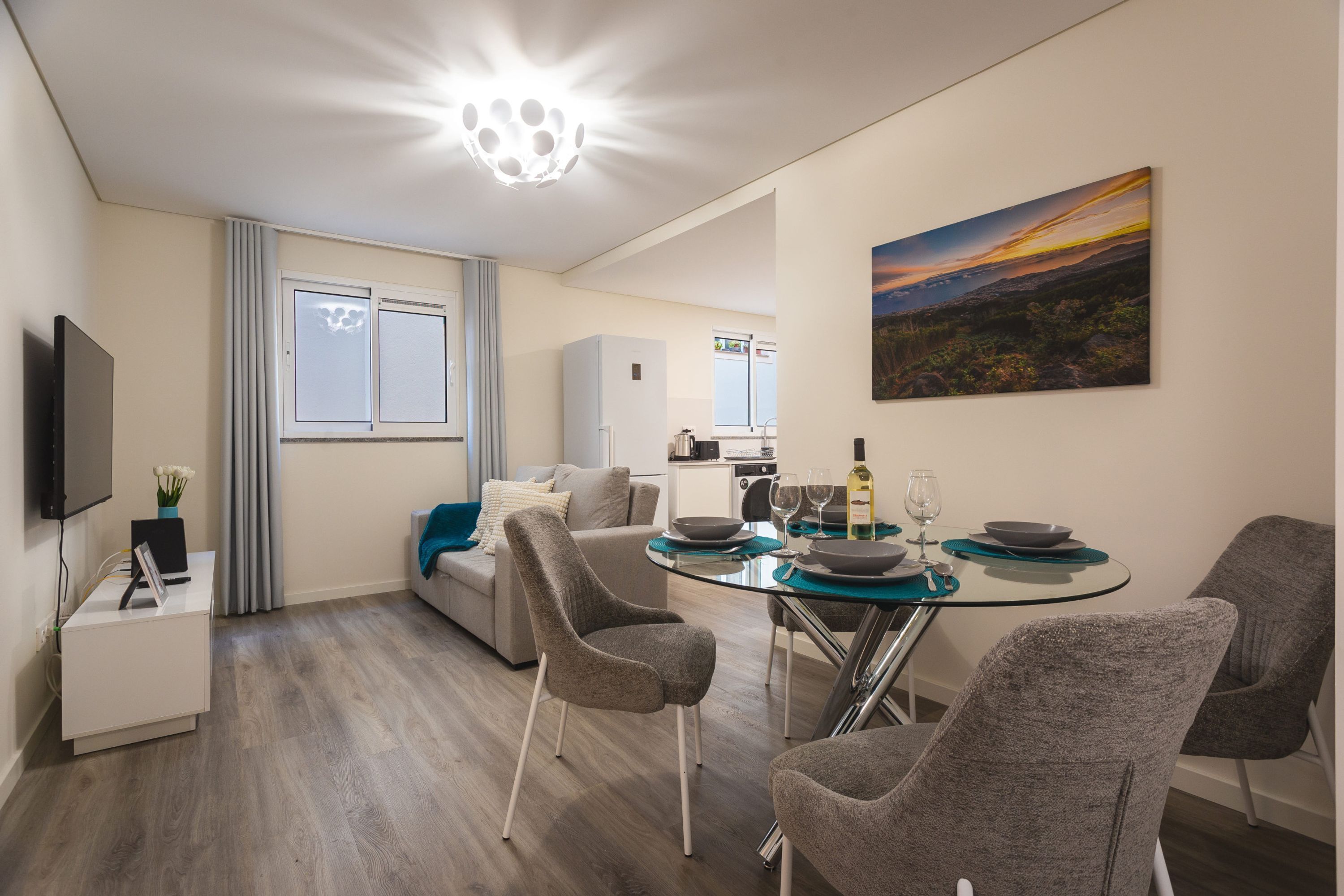 Een moderne woonkamer met een glazen eettafel, stoffeerde grijze stoelen, een grijze bank, een flatscreen televisie en een schilderij aan de muur.