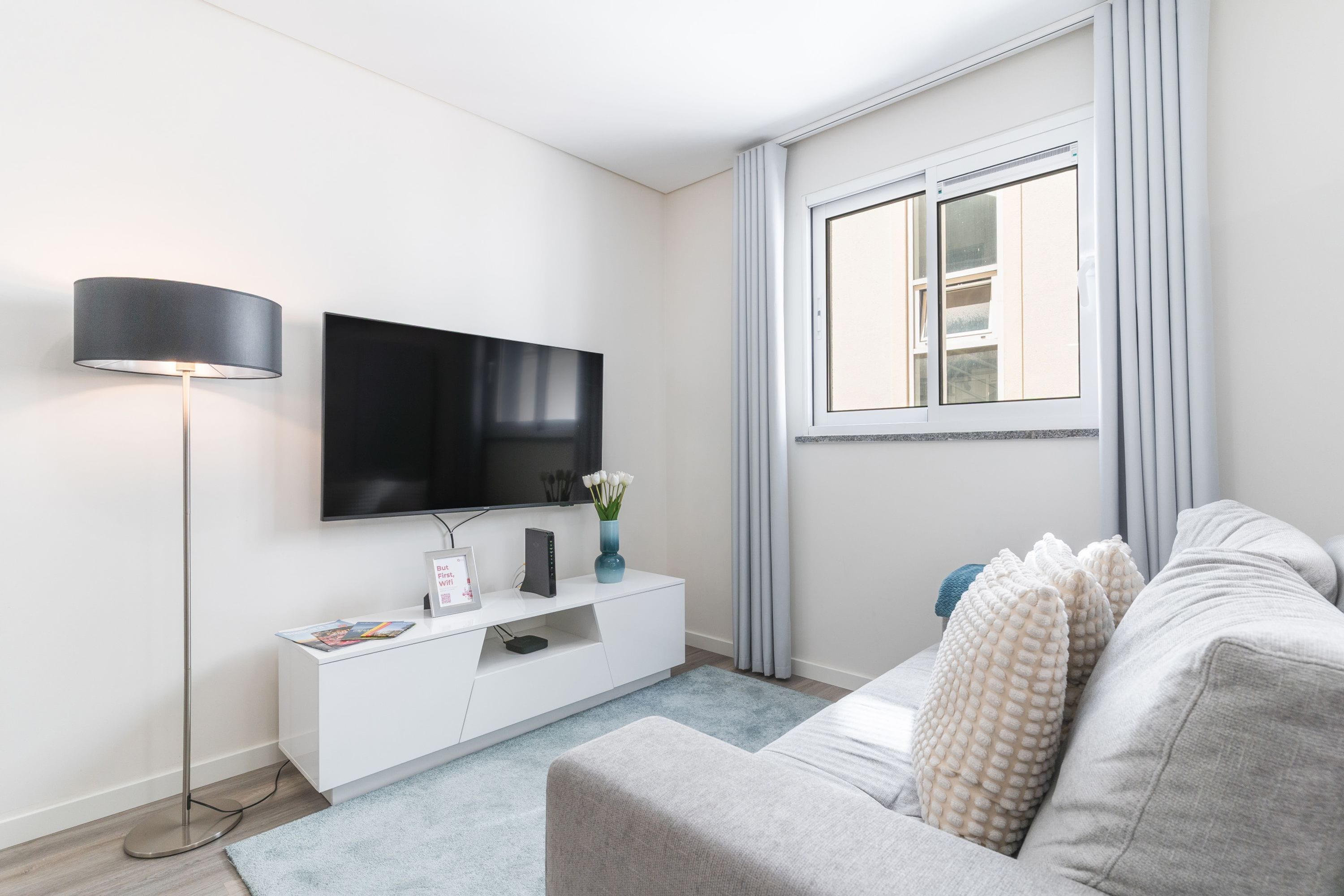 Ein modernes Wohnzimmer mit einem Flachbildfernseher, einer weißen Konsole, einem grauen Sofa, einer Stehlampe und einem Fenster mit Vorhängen.