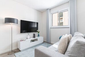 A modern living room with a flat-screen TV, a white console table, a grey sofa, a floor lamp, and a window with curtains. - Funchal Essence by Atlantic Holiday (Funchal)