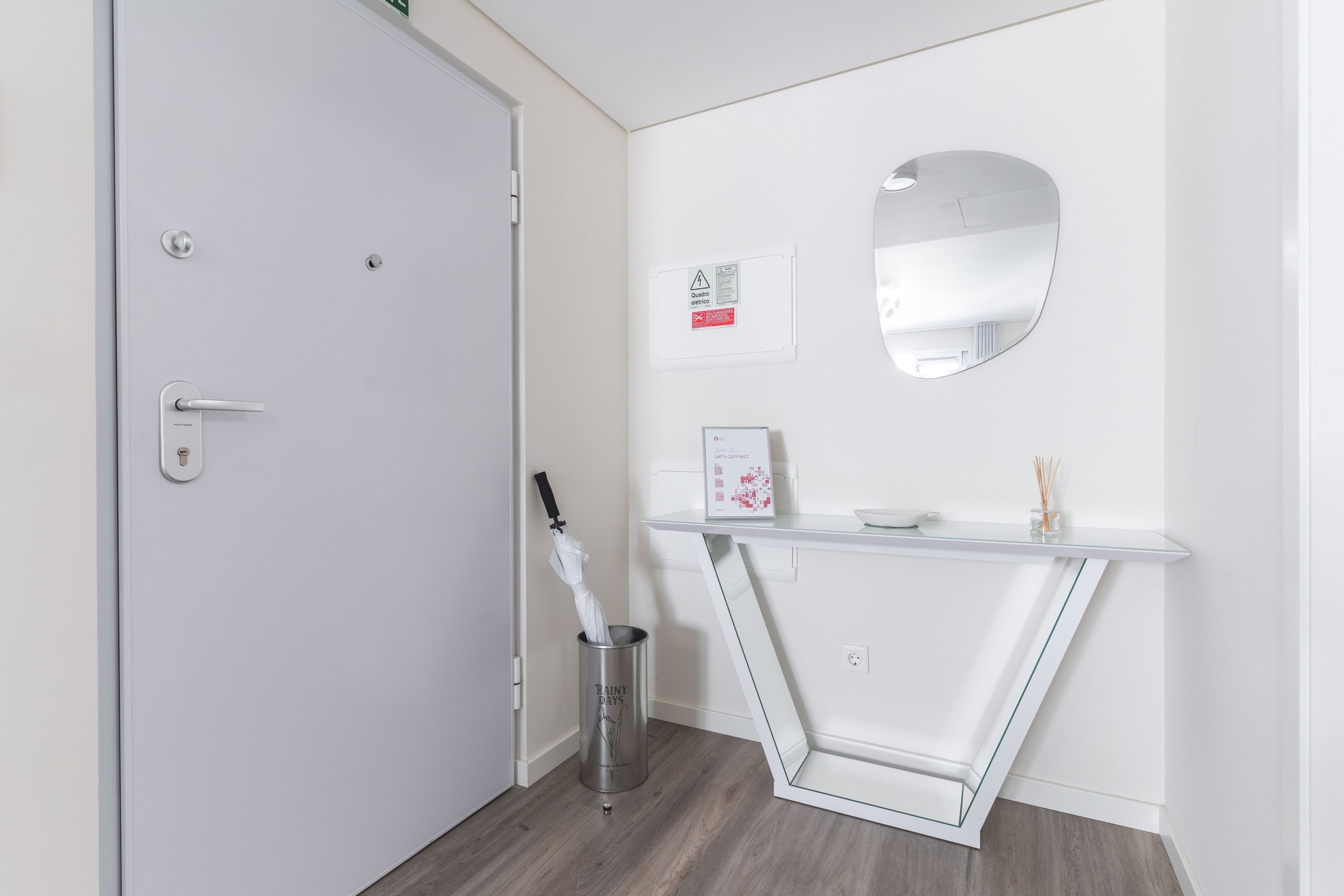 Un baño moderno con una puerta blanca, un espejo, un pequeño cubo de basura y una repisa de vidrio con un cuadro enmarco y un cuenco.