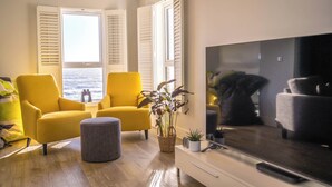 A modern living room with a large TV, yellow armchairs, a grey ottoman, and a view of the sea through open windows. - Wight Stone Luxury Townhouse - Breathtaking Views  (Sandown)