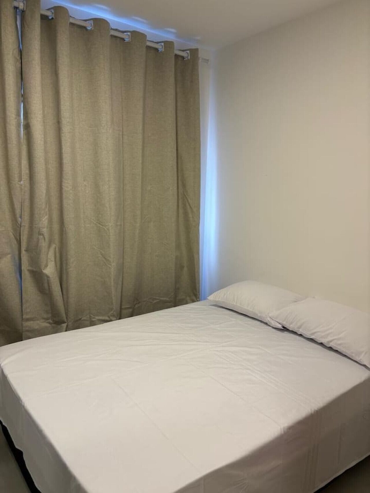 Habitación con una cama individual, cubierta con una colcha blanca sencilla y dos almohadas. Una cortina beige cubre parcialmente una ventana, y las paredes son de color blanco.