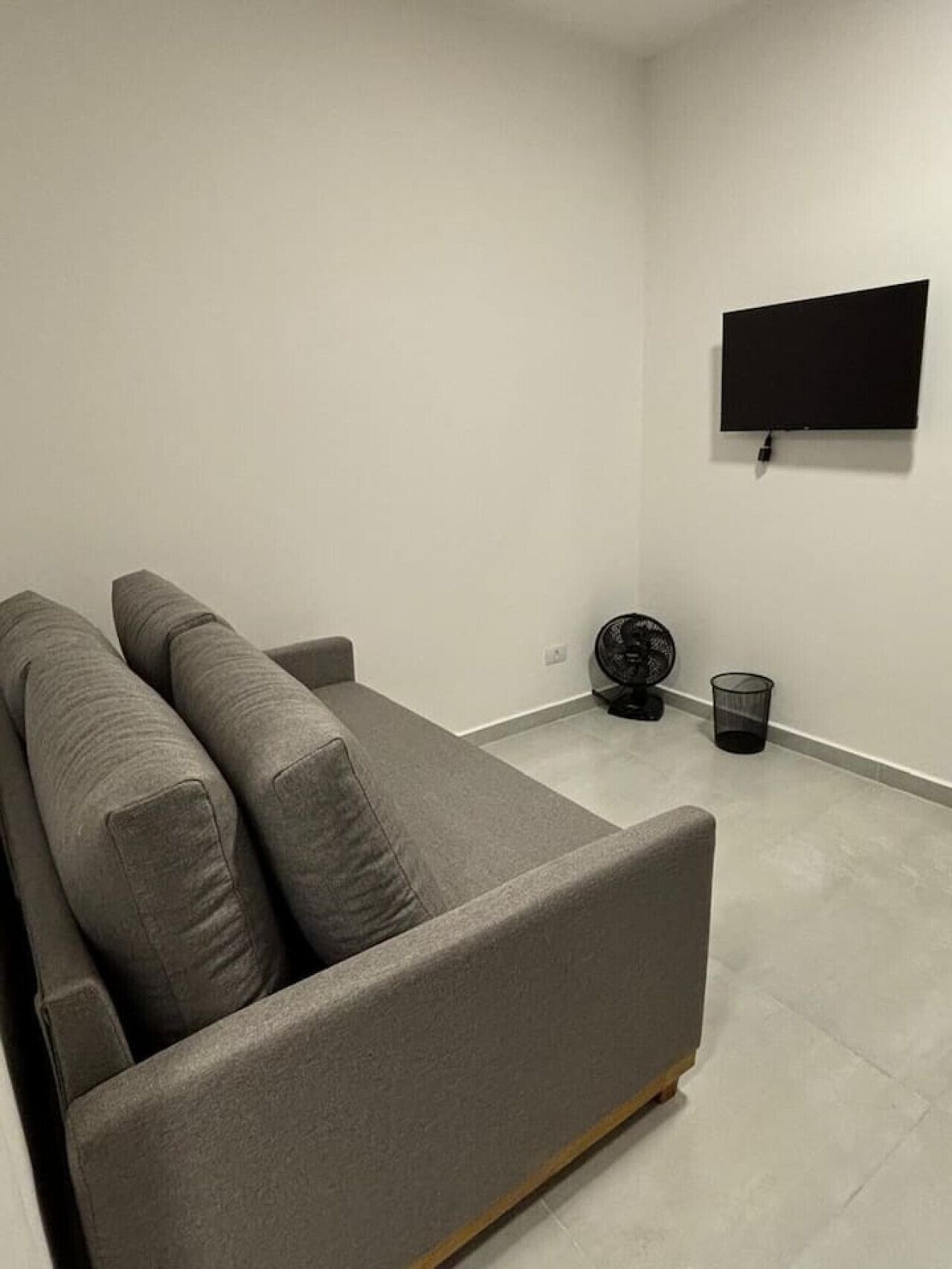 Ein minimalistisches Wohnzimmer mit grauem Sofa, einem an der Wand montierten Fernseher, einem Standventilator und einem Mülleimer.