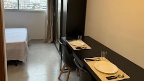 A small, modern room with a dining table set for two, a bed, and a large window offering a city view. - Studio 612 - Tucuruvi - 20min do Anhembi (São Paulo)