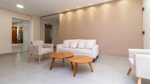 A modern hotel lobby with a white sofa, two wooden coffee tables, and a glass partition. - Studio 612 - Tucuruvi - 20min do Anhembi (São Paulo)