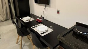A modern kitchen with a black countertop, a dining table with chairs, a TV mounted on the wall, and a small island with a sink. - Studio novo 405 - metrô Santana - sem Wi-Fi (São Paulo)